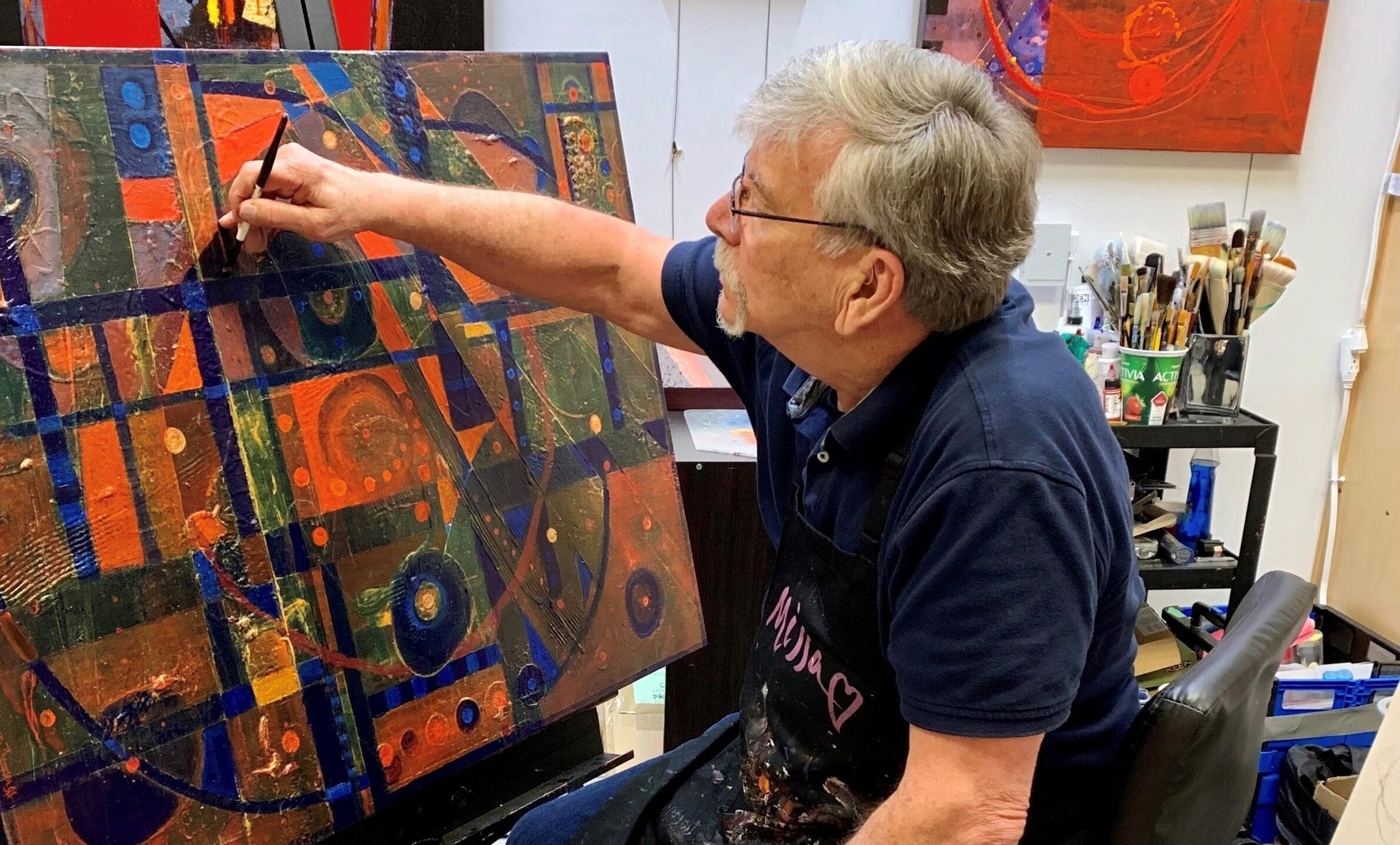 HB Barry Strasbourg-Thompson at work in his studio, brush in hand, painting into a large grid-and-circle canvas in orange, green and cobalt.